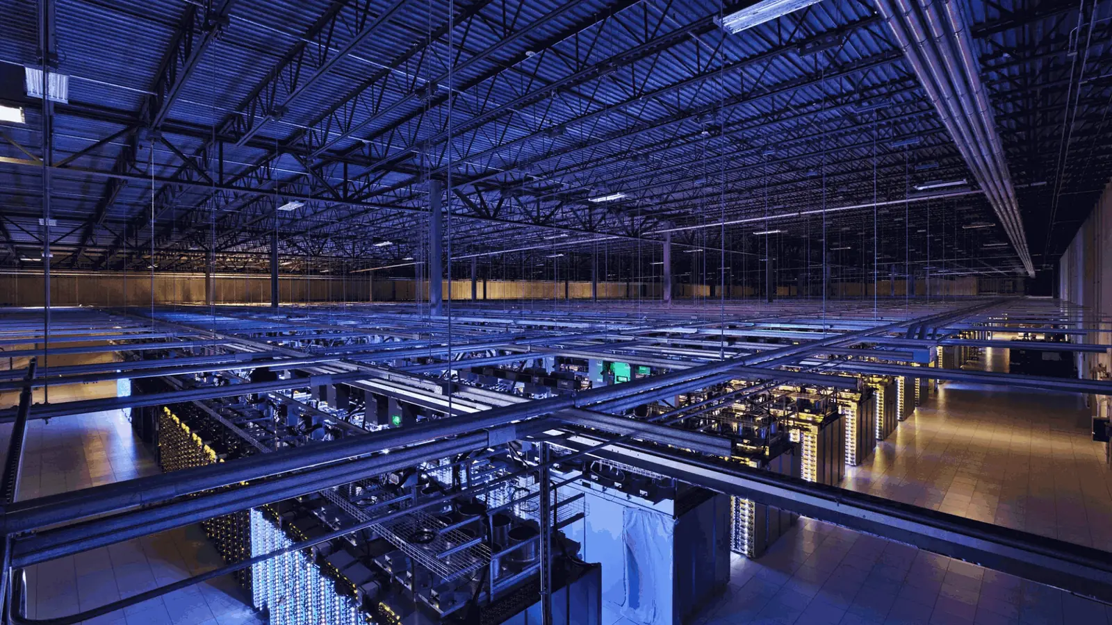 A data hall seen across its overhead cable trays, with rows of server racks below in blue light